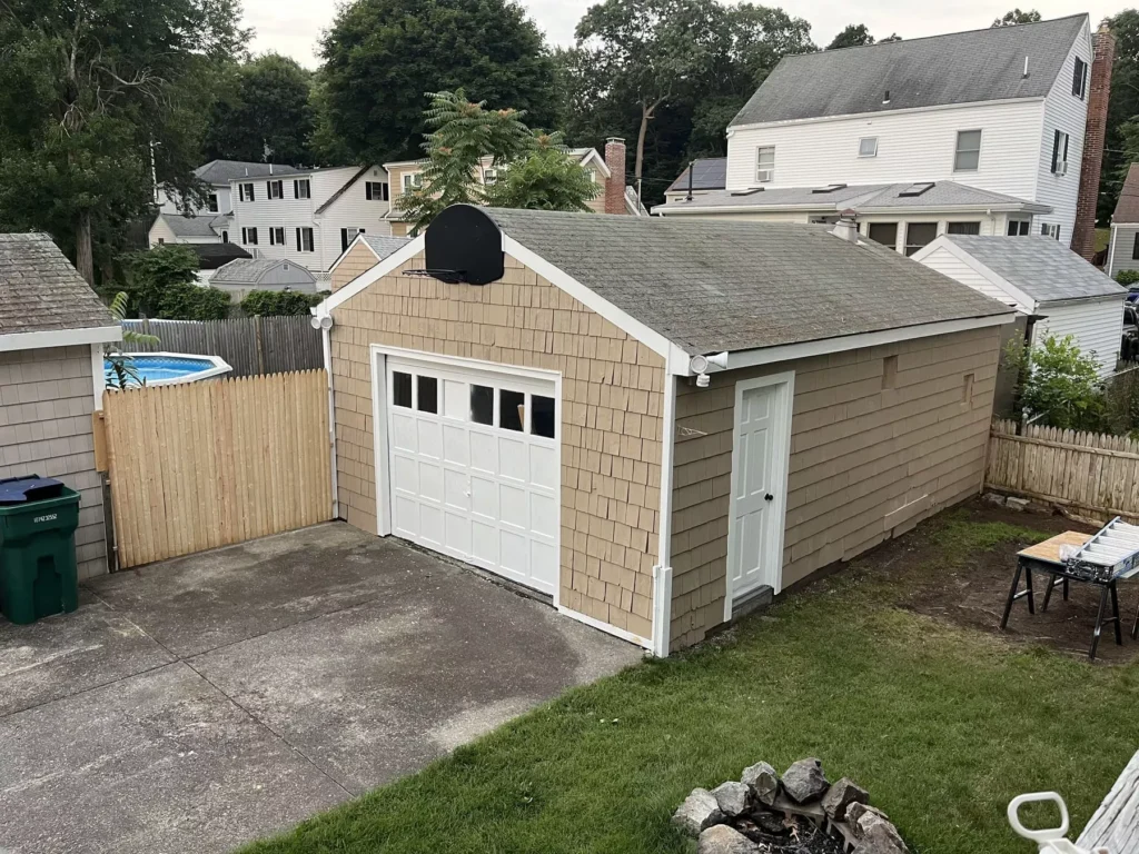 Tan shingle detached garage, a potential ADU, with a white garage door, basketball hoop, and a grassy backyard.