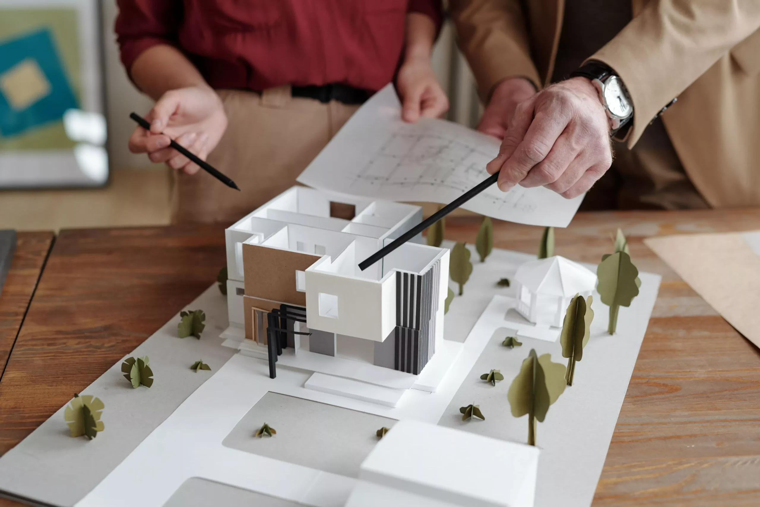 Two pairs of hands, one holding blueprints, point with pencils at a modern white and brown house model with paper trees on... Two pairs of hands, one holding blueprints, point with pencils at a modern white and brown house model with paper trees on...