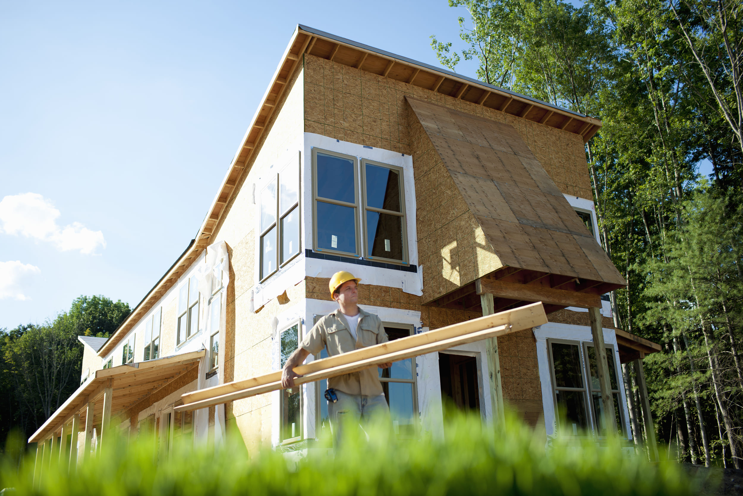 Construction worker in yellow hard hat carrying wood planks in front of a house under construction with green trees and bl... Construction worker in yellow hard hat carrying wood planks in front of a house under construction with green trees and bl...