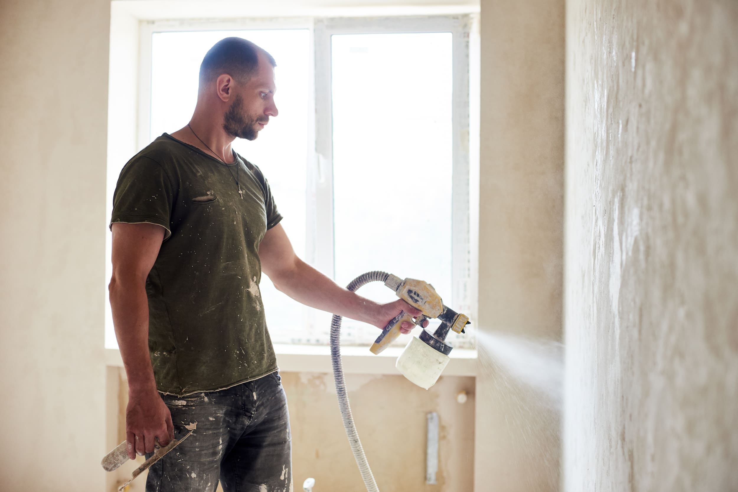 Bearded man in dirty green shirt painting a textured beige wall with a paint sprayer during home renovation. Bearded man in dirty green shirt painting a textured beige wall with a paint sprayer during home renovation.